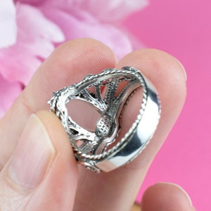 May include: A silver ring with an intricate filigree design. The ring features a detailed openwork pattern and a polished band. The ring is held against a pink background, showcasing its ornate details and craftsmanship.