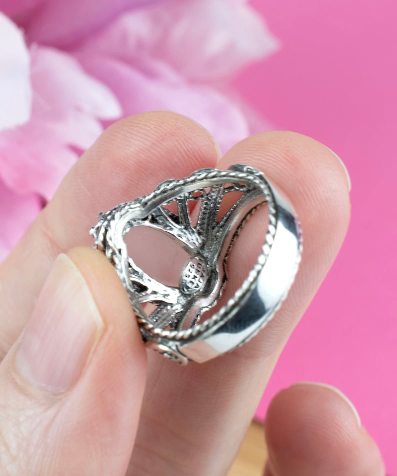 May include: A silver ring with an intricate filigree design. The ring features a detailed openwork pattern and a polished band. The ring is held against a pink background, showcasing its ornate details and craftsmanship.