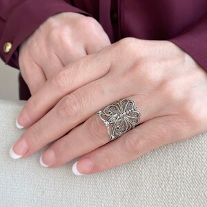 May include: A silver filigree butterfly ring with intricate details. The ring is worn on a finger with a white nail polish manicure.