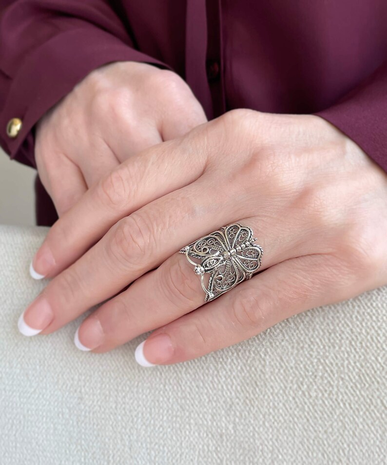 May include: A silver filigree butterfly ring with intricate details. The ring is worn on a finger with a white nail polish manicure.