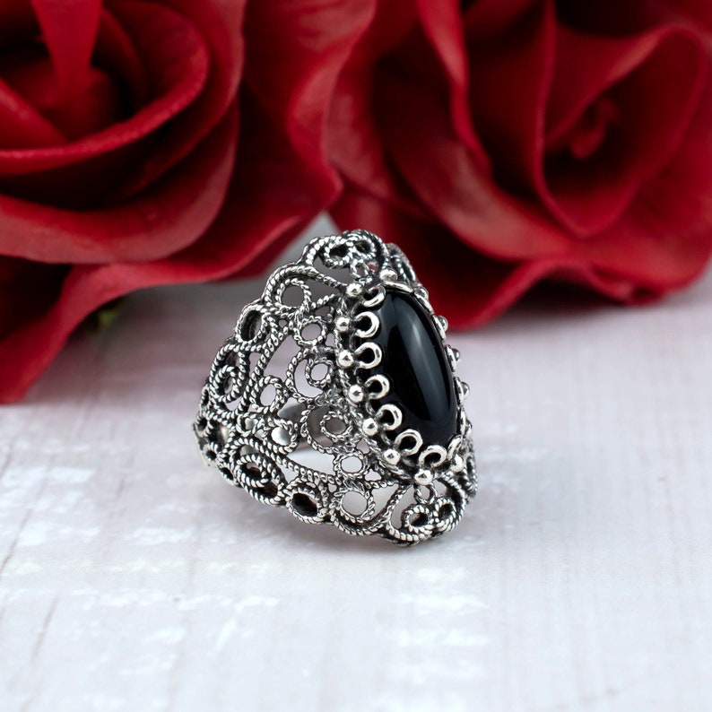 May include: A silver filigree ring with a black oval gemstone. The ring is set against a background of red roses.