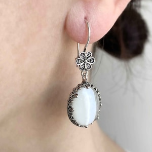 May include: Silver dangle earrings with a white oval-shaped stone and a flower-shaped charm.