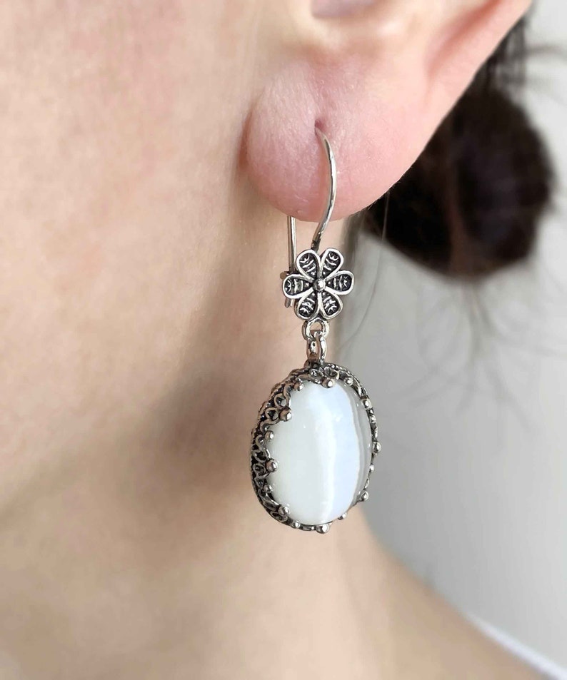 May include: Silver dangle earrings with a white oval-shaped stone and a flower-shaped charm.