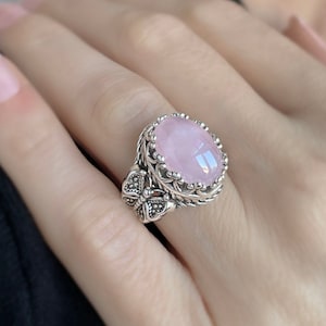 May include: A silver ring with a large oval pink gemstone set in a detailed filigree design. The ring features a butterfly design on the side.