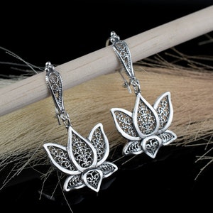 May include: A pair of silver filigree earrings with a lotus flower design. The earrings are hanging from a wooden surface.