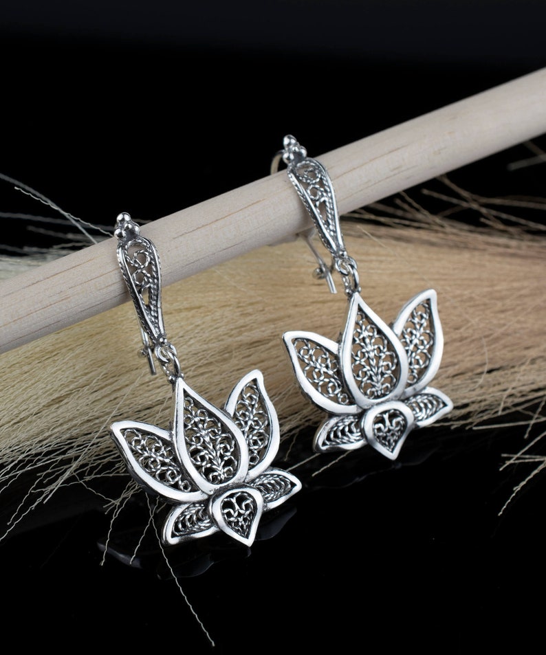 May include: A pair of silver filigree earrings with a lotus flower design. The earrings are hanging from a wooden surface.