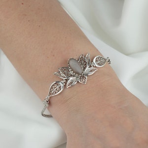 May include: A silver bracelet with a delicate filigree design featuring a gray stone in the center. The bracelet is shaped like a lotus flower.