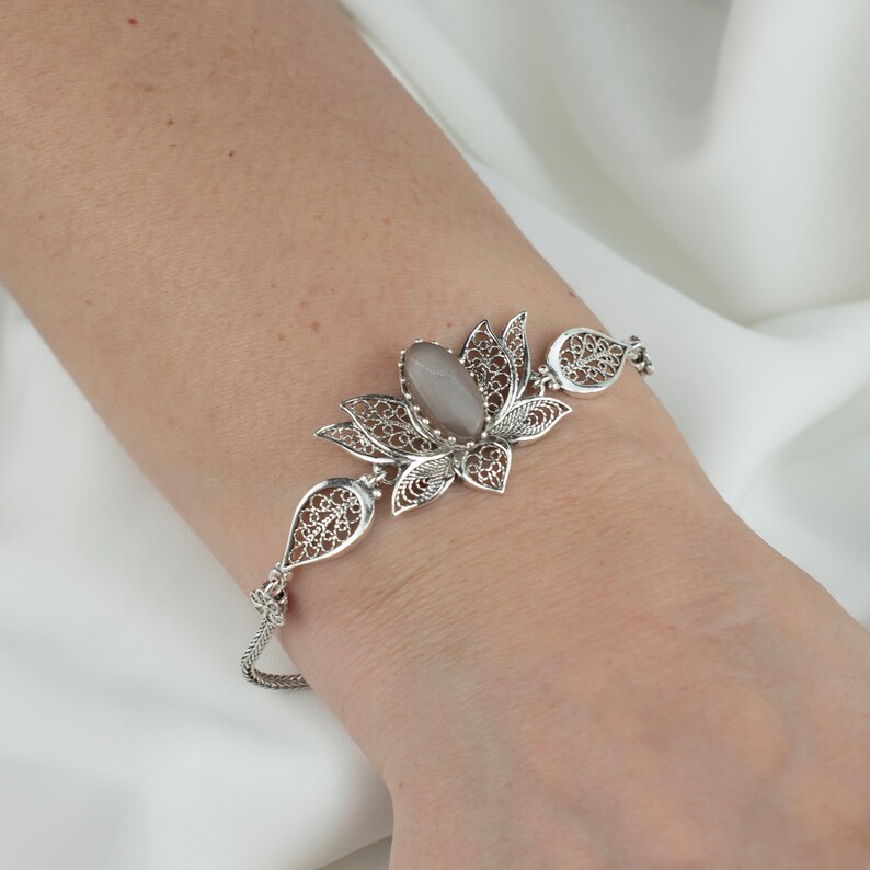 May include: A silver bracelet with a delicate filigree design featuring a gray stone in the center. The bracelet is shaped like a lotus flower.