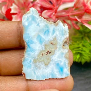 May include: A flat, irregular piece of polished Larimar stone featuring a marbled pattern of sky blue and white with a central patch of tan and brown inclusions. The stone has a smooth, polished surface and a jagged, natural-looking top edge.