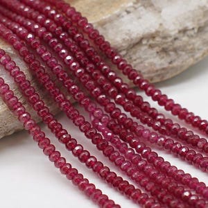 May include: Multiple strands of small, faceted, deep red gemstone beads arranged in parallel lines. Each bead features a uniform round shape with a reflective, polished surface that catches the light, creating a consistent texture across the collection.