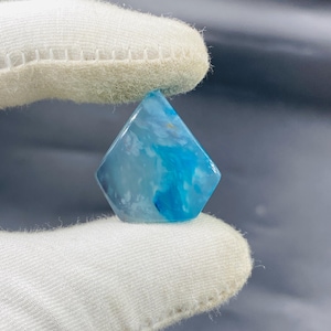 Könnte beinhalten: Ein rautenförmiger Edelstein in Blau- und Weißtönen. Der Stein hat ein wolkiges Aussehen mit Bereichen in Hell- und Dunkelblau. Er wird von einer behandschuhten Hand gehalten.