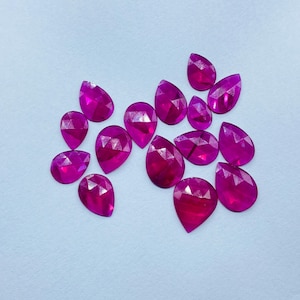 May include: A collection of faceted, teardrop-shaped, fuchsia-coloured gemstones. The gems are arranged on a light blue surface, showcasing their cut facets and vibrant colour. The gems vary in size.