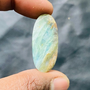May include: An oval gemstone with a polished surface. The stone displays a mix of light green, blue, and touches of brown and yellow. The gemstone is held between two fingers, highlighting its size and shape.