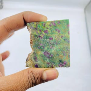 May include: A rectangular gemstone with a rough edge. The stone has a green base colour with red and purple spots. The edges are a mix of brown and tan. The stone is held in a hand.