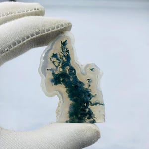 May include: A polished, irregularly shaped gemstone with a white and translucent base. The stone features striking inclusions of deep green, creating a natural, artistic pattern. The gemstone is held by a gloved hand.