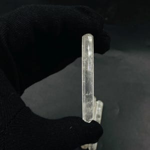 Puede incluir: Un cristal claro y alargado con forma rectangular facetada. El cristal se sostiene sobre un fondo oscuro, mostrando su transparencia y texturas internas. Es un espécimen mineral natural.