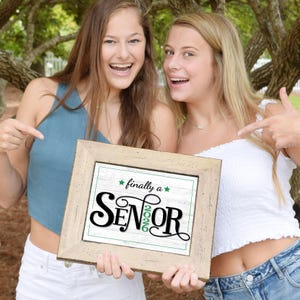 Two teen girls holding a framed First day of senior year sign or photo prop with green accents that says "Finally a Senior" and "2026"