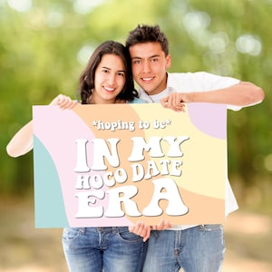 Puede incluir: Una pareja sonriente sostiene un cartel con el texto blanco "*hoping to be* IN MY HOCO DATE ERA". El cartel tiene un diseño pastel de rosa, azul y amarillo. El fondo es verde borroso.