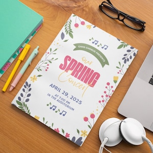 May include: A white concert flyer with the text "Our Spring Concert" in pink and yellow, surrounded by floral illustrations. The flyer includes the date, time, and location of the event. A pair of glasses, headphones, and pens are on the wooden surface.
