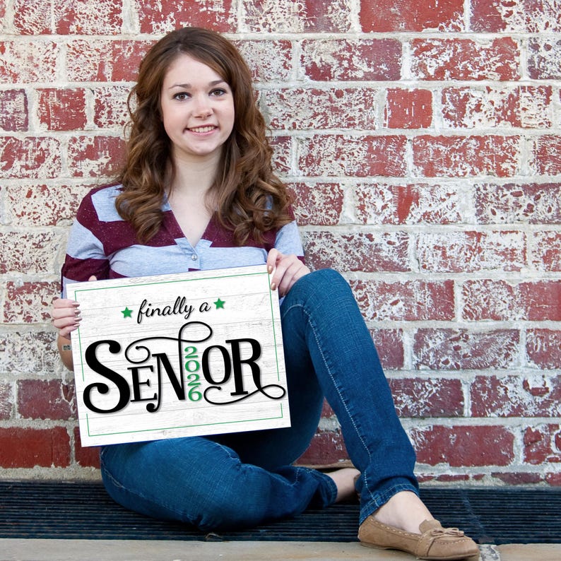 Teen girl sitting against a brick wall holding a first day of senior year sign or photo prop with green accents that says "Finally a Senior" and "2026"