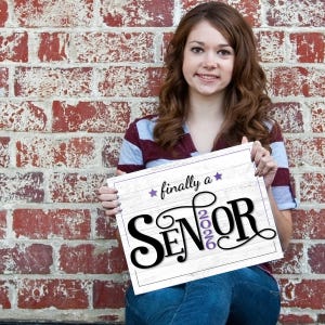 Teen girl sitting against a brick wall holding a purple "Finally a Senior" sign for the class of 2026