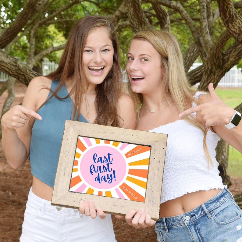 Two teen girls holding and pointing to a Last First Day sign or photo prop for the class of 2026, sunrise design in pink and orange