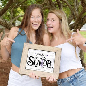 Two happy teen girls holding a framed Purple first day of senior year sign for the class of 2026