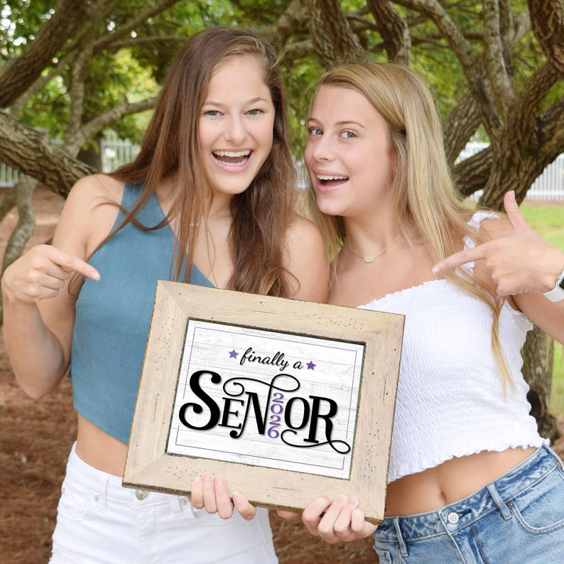 Two happy teen girls holding a framed Purple first day of senior year sign for the class of 2026