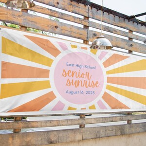 Op de afbeelding: Een rechthoekige banner met een zonnestraalpatroon in tinten geel, oranje en roze. Een grote roze cirkel in het midden toont de tekst East High School, senior sunrise en August 16, 2025, in blauwe en oranje schrijfletters en drukletters.