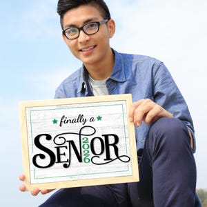 Teen boy holding a framed First day of senior year sign or photo prop with green accents that says "Finally a Senior" and "2026"