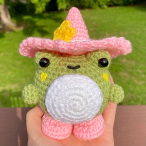 Puede incluir: Una muñeca de rana de ganchillo hecha a mano con un sombrero de bruja rosa adornado con una estrella amarilla. La rana es verde con vientre blanco, ojos negros y pies rosas. La muñeca está en una mano.