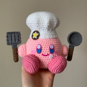 Könnte beinhalten: Ein rosa gehäkeltes Kirby Plüschtier, das einen weißen gehäkelten Kochhut mit einem gelben Stern trägt. Das Plüschtier hält zwei gehäkelte Spatel, einen grauen und einen braunen.