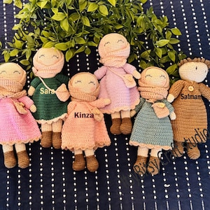 May include: A collection of handmade crochet dolls in various colors, including brown, pink, green, and blue. Each doll has a unique outfit and is labeled with a name. The dolls are arranged on a dark blue surface with white polka dots.