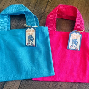 May include: Two small, fabric tote bags with handles. One bag is blue and the other is pink. Each bag has a tag attached with the text "Thank you for joining [name]'s party!" and a cartoon image of Sonic the Hedgehog.