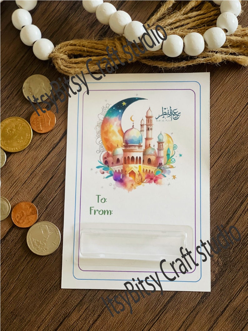 May include: A white card with a colorful illustration of a mosque and crescent moon. The card has a clear plastic insert for a gift card or money. The text on the card reads "Eid al-Fitr".