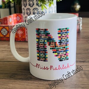 May include: White ceramic mug with a colorful crayon letter "M" and the text "Miss Nahlilah" in red. The mug is on a wooden surface.