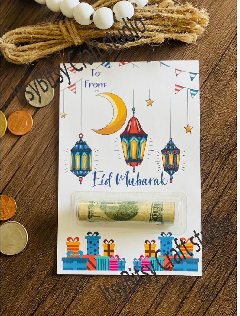 May include: A white card with a crescent moon and lanterns, the text "Eid Mubarak" and a rolled up dollar bill inside a clear plastic sleeve. The card is decorated with colorful gift boxes and red, white, and blue bunting.