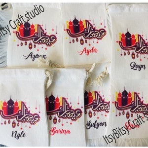 May include: Five white canvas drawstring bags with colorful Arabic calligraphy and a mosque design. Each bag has a different name printed on it: Azan, Aydn, Qayn, Nyle, and Sarina.