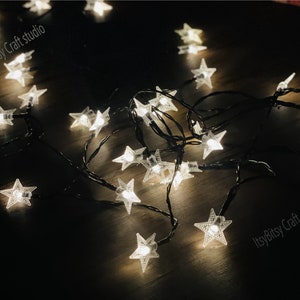 May include: A string of white star-shaped fairy lights with a black wire. The lights are scattered on a wooden surface.