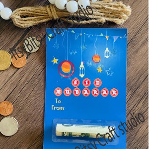 May include: Blue card with a colorful illustration of stars, crescent moons, and lanterns. The card says "Eid Mubarak" in red circles and has a slot for cash. The card is on a wooden surface with white beads and a brown tassel.