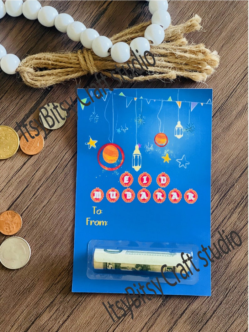 May include: Blue card with a colorful illustration of stars, crescent moons, and lanterns. The card says "Eid Mubarak" in red circles and has a slot for cash. The card is on a wooden surface with white beads and a brown tassel.