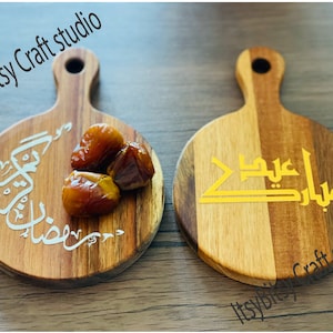May include: Two round wooden serving trays with handles. One tray has a white Arabic inscription and dates on it. The other tray has a gold Arabic inscription. Both trays have the text "Itsy Bitsy Craft Studio" on them.
