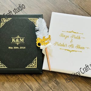 Puede incluir: Dos libros de visitas de boda, uno negro y otro blanco, con letras y detalles dorados. El libro negro tiene "K&M" y la fecha "May 30th, 2014". El libro blanco tiene nombres en escritura dorada. Un bolígrafo decorativo con una pluma está entre los libros.