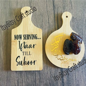 May include: Two wooden serving trays with handles. One tray is rectangular and has the text "NOW SERVING... Iftar TILL Suhoor". The other tray is round and has Arabic calligraphy in gold. Both trays have dates on them.
