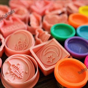 Op de afbeelding: Een set roze plastic koekjesvormen met islamitische ontwerpen, waaronder een halve maan, een ster, een moskee en een lantaarn. De vormen zijn gerangschikt op een houten oppervlak met kleurrijke playdough-containers op de achtergrond. De tekst "ItsyBitsy Craft studio" is zichtbaar op twee van de containers.