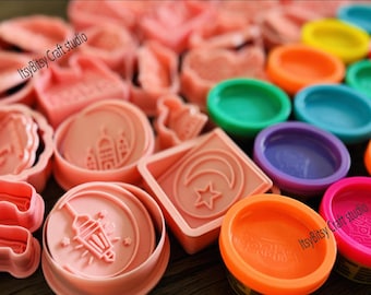 May include: A set of pink plastic cookie cutters featuring Islamic designs, including a crescent moon, a star, a mosque, and a lantern. The cutters are arranged on a wooden surface with colorful play dough containers in the background. The text "ItsyBitsy Craft studio" is visible on two of the containers.