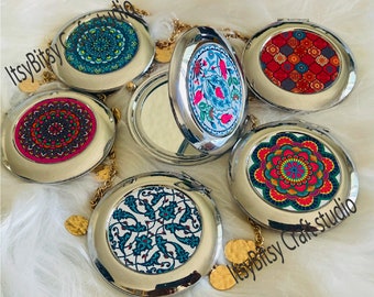 May include: Six silver compact mirrors with different colorful patterned designs. Each mirror has a gold chain with a decorative coin charm. The mirrors are on a white fluffy surface. The text "Itsy Bitsy Craft studio" is visible in the bottom right corner.