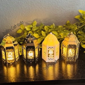 Op de afbeelding: Vier decoratieve lantaarns in goud, zwart en wit, elk met ingewikkelde uitgesneden ontwerpen en een warme, uitnodigende gloed. De lantaarns zijn gerangschikt op een donker oppervlak met groene bladeren op de achtergrond. De tekst "ItsyBitsy Craft studio" is zichtbaar.