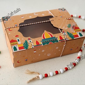 May include: A brown cardboard gift box with a clear window and a decorative design featuring colourful buildings, moons, and stars. The box is tied with twine and includes a tag. The words "Itsy Bitsy Craft studio" are printed on the box.