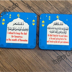 May include: Two blue square coasters with a crescent moon and lanterns. The coasters feature Arabic text and English translations. The text on the left coaster reads "I intend to keep the fast for tomorrow in the month of Ramadan." The text on the right coaster reads "Oh Allah! I fasted for You and I believe in You and I put my trust in You and I break my fast with your sustenance."